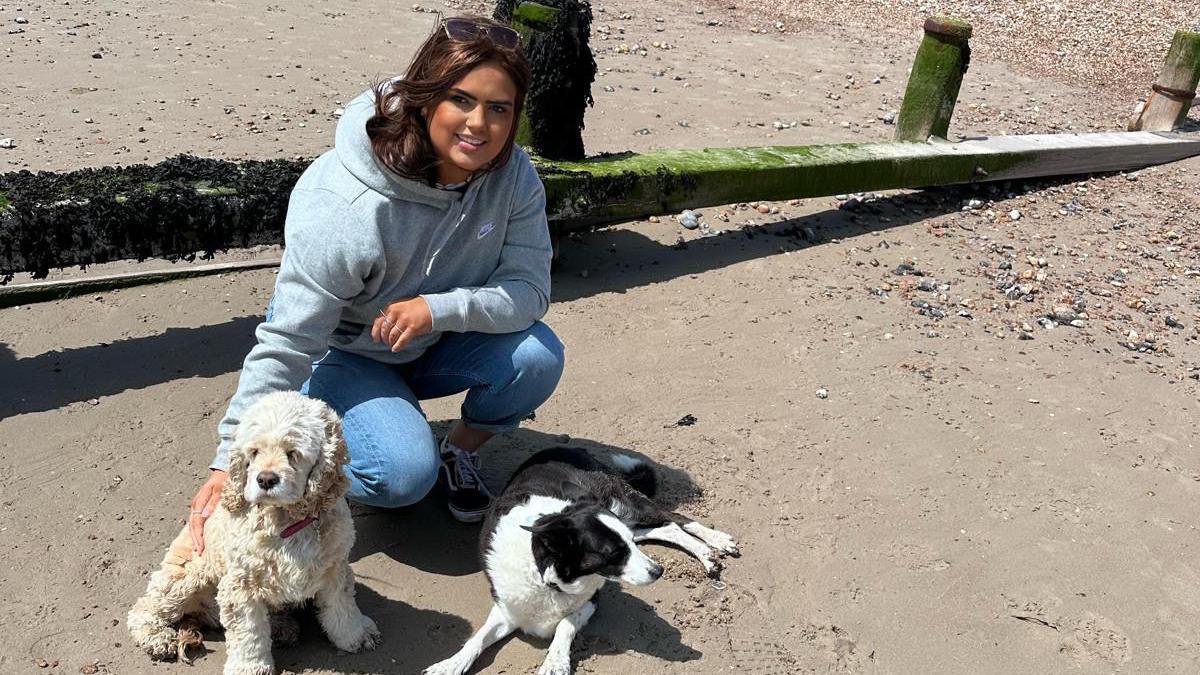 Hannah is crouching on the floor on a beach, with two dogs near her. She is looking at the camera and smiling.