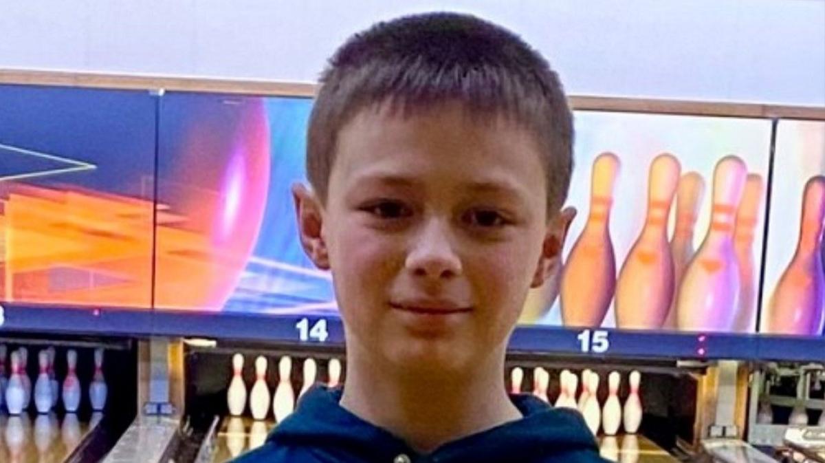 A close-up photo of a boy looking straight at the camera. He has brown hair and he is wearing a blue top. He is in a 10-pin bowling alley, with lanes and sets of pins visible behind him.
