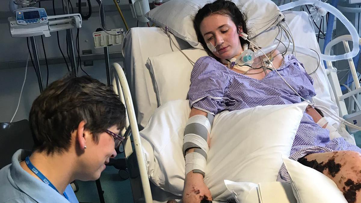 Becca Heritage laying in a hospital bed, wearing a purple striped hospital down. She is attached to lots of wires and medical equipment, including a large tube attached to her throat. Her legs and arms have black patches on them. A female healthcare professional wearing an NHS lanyard is crouched by her bedside.