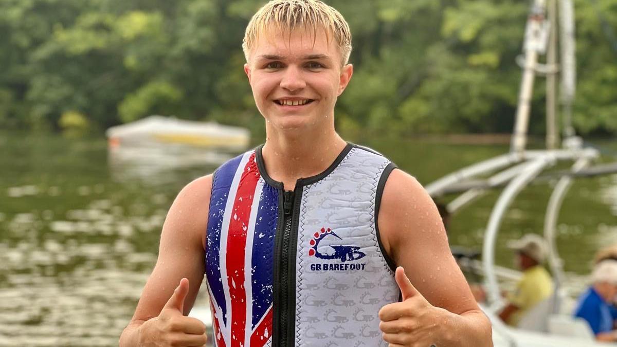 A boy with blonde hair is standing by the water giving both thumbs up. He is wearing a neoprene vest in the British flag colours.