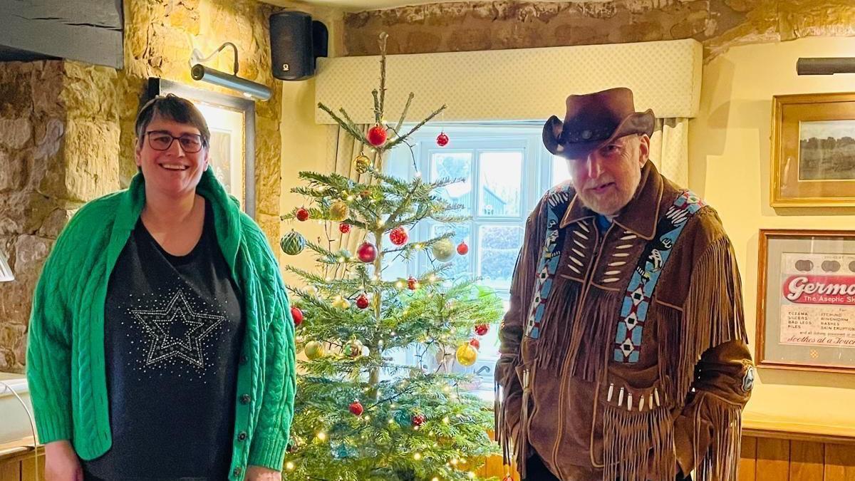 A woman in a green jumper and a man in a cowboy outfit smile in front of a Christmas tree.
