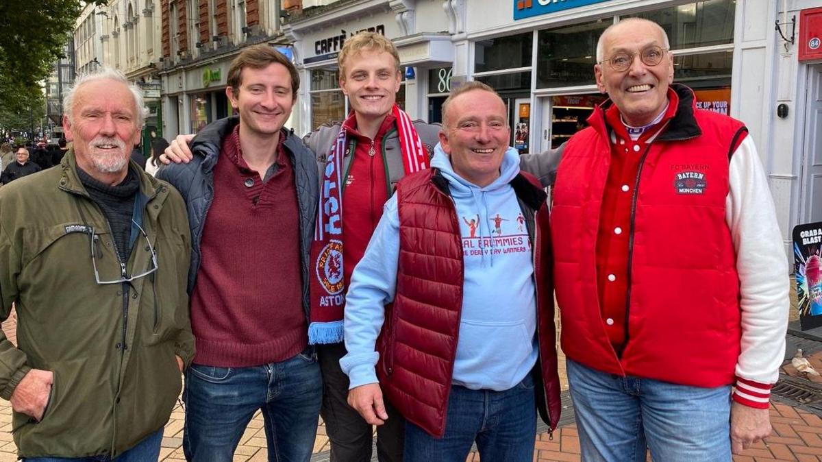 Five men are standing in a row and smiling. They are all wearing jackets and standing on a cobbled street in Birmingham city centre 