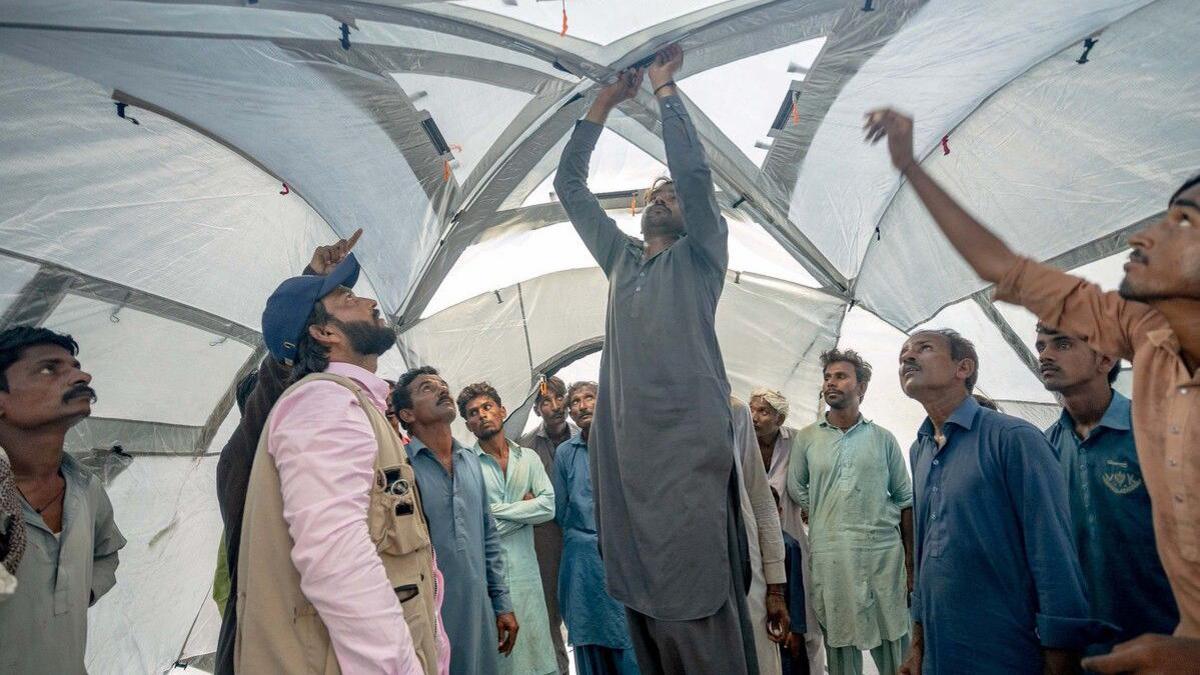 A group of men stand inside a large tent. They are looking at one man in the centre of the image who appears to be putting part of the tent together. 