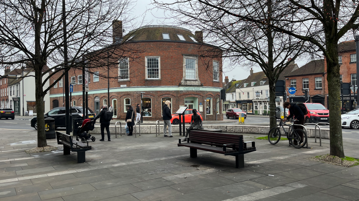 A shot of a town square in Chichester with bences and trees around a paved aread with a large municipal building in the background 