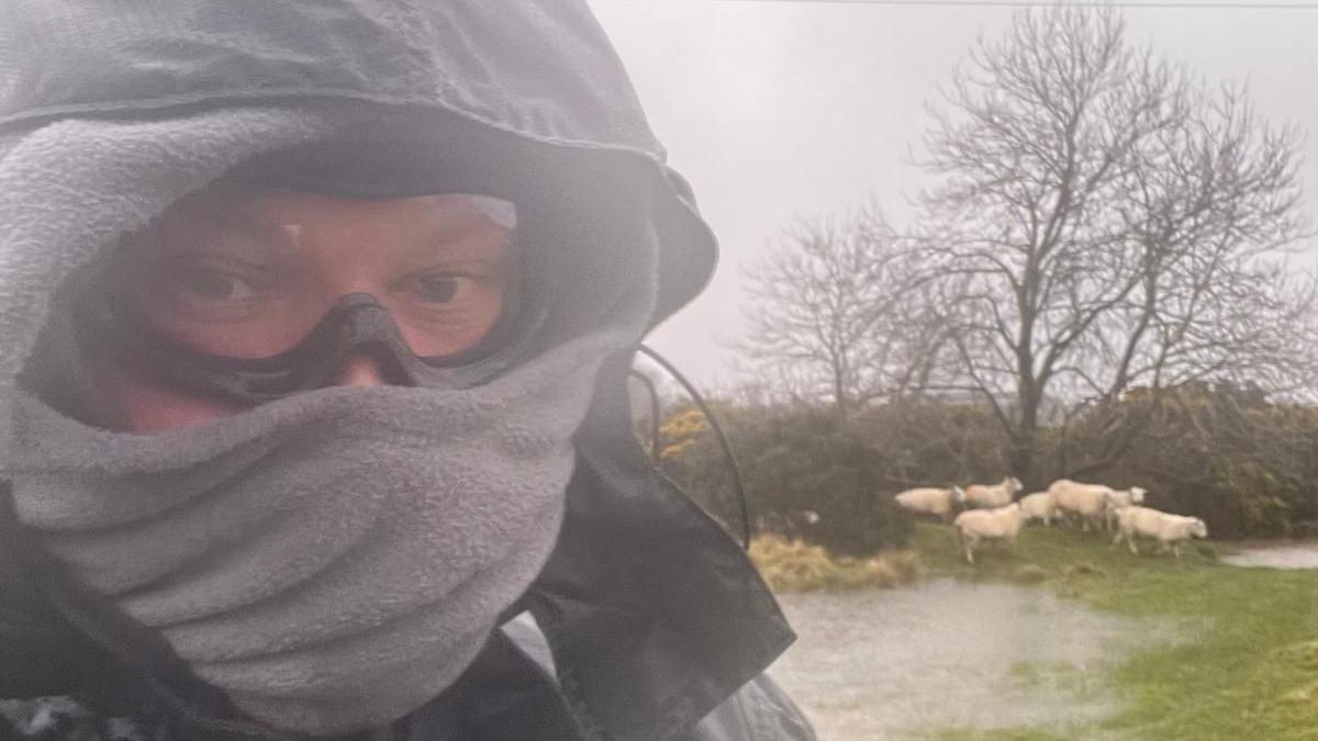 A picture of a farmer wrapped up in pouring rain with a waterlogged field behind 