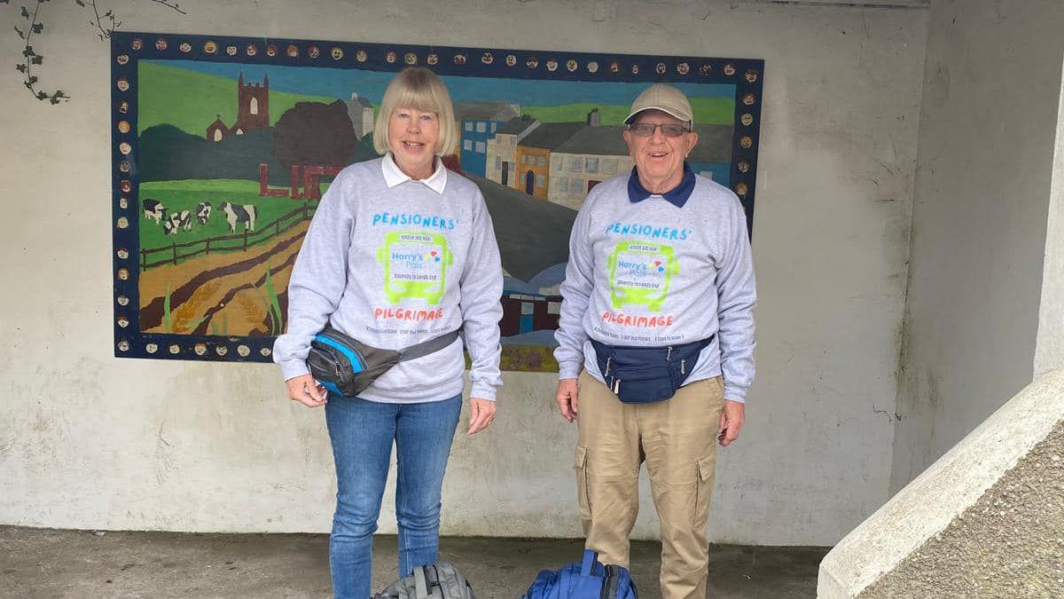 Mr and Mrs Banham wearing their grey Pilgrimage sweatshirts and belt packs. Mr Banham is wearing a brown cap. They are standing in front of a mural showing a village scene with a church, houses, cows and a river.