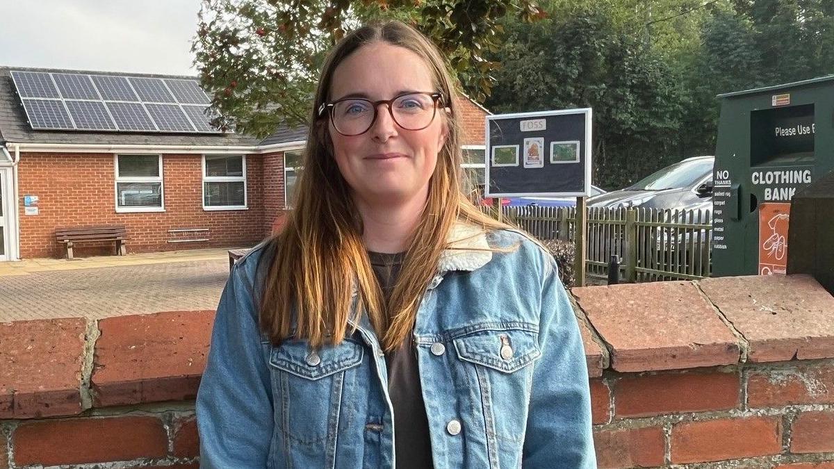 Cassie Holborow, with long copper brown hair, is wearing a blue demin jacket and dark spectacles. She is pictured in front of Scampton C of E Primary School.  