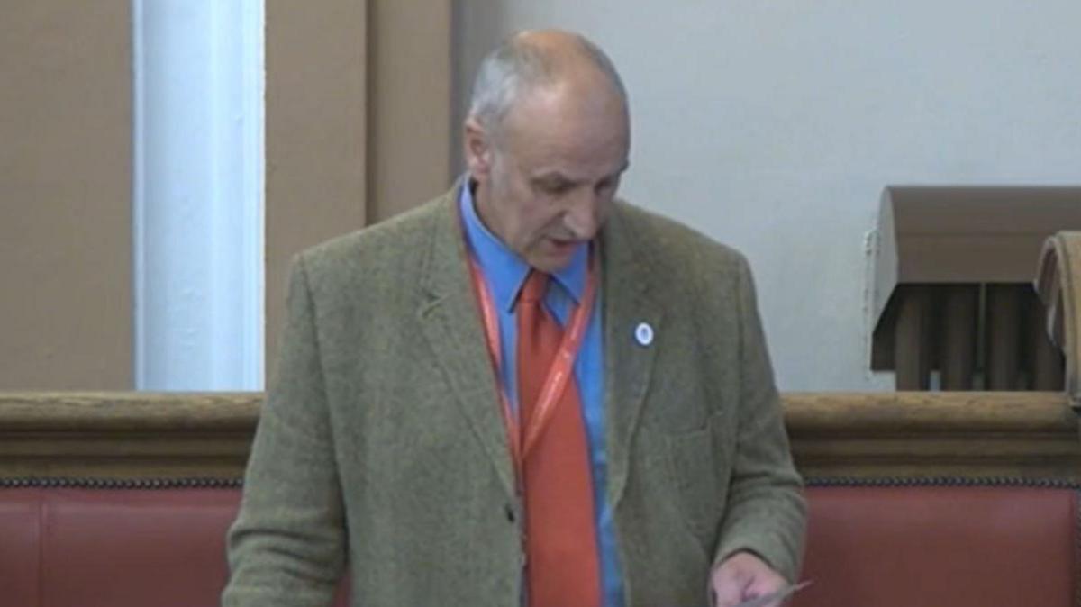 David Mountford, a man wearing a blue shirt and red tie with a green jacket, is standing in a council chamber. He is holding a piece of paper from which he appears to be reading.
