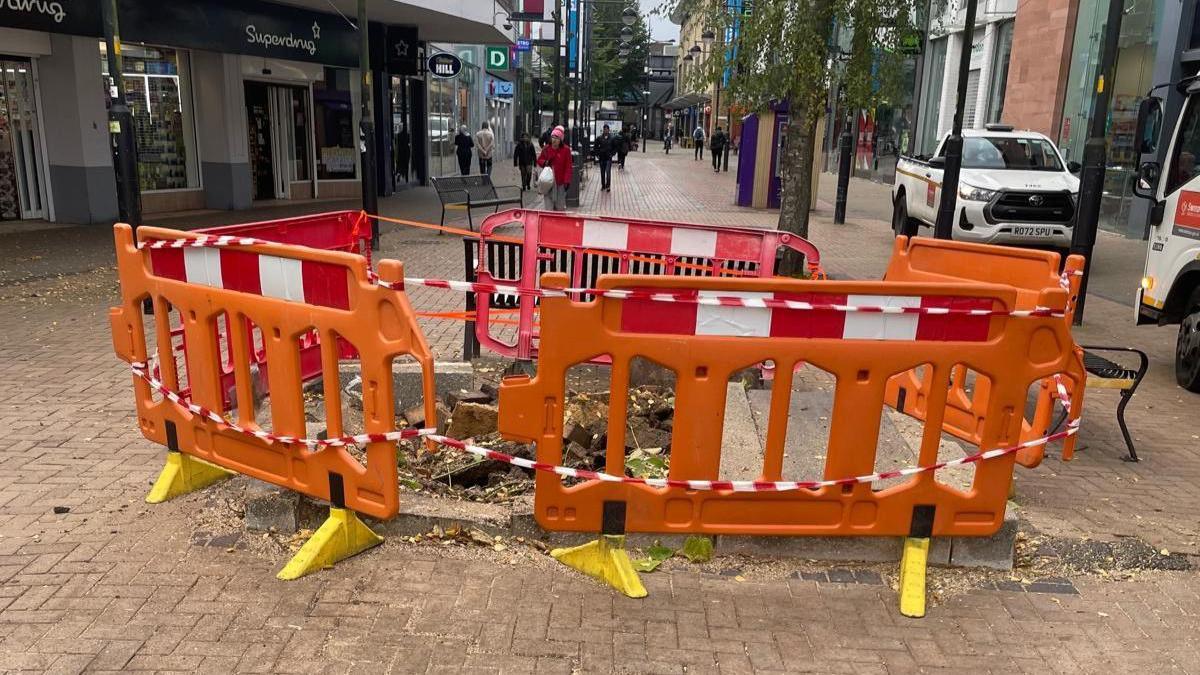 Orange plastic barriers and red and white tape cordoning off an area of The Parade where the tree once stood. The concrete base it was in has been ripped up and the stump of the tree is still visible. There are shops lining the street and people walking in the background.