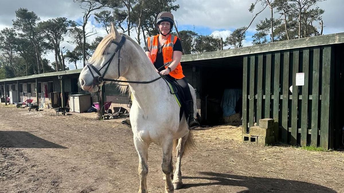 A picture of Laura Hunter sitting on a grey horse wearing a high vis jacket 