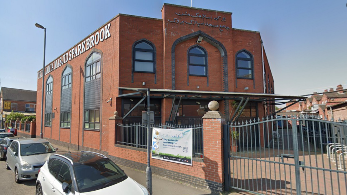 A large two-storey red brick building on a sunny day. The building has a metal gate at its entrance and silver Arabic writing across the front of the building.
