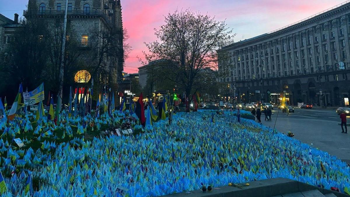 Hundreds of blue and yellow flags fill a square with bigger Ukrainian flags to the left with a street and people walking on it to the right