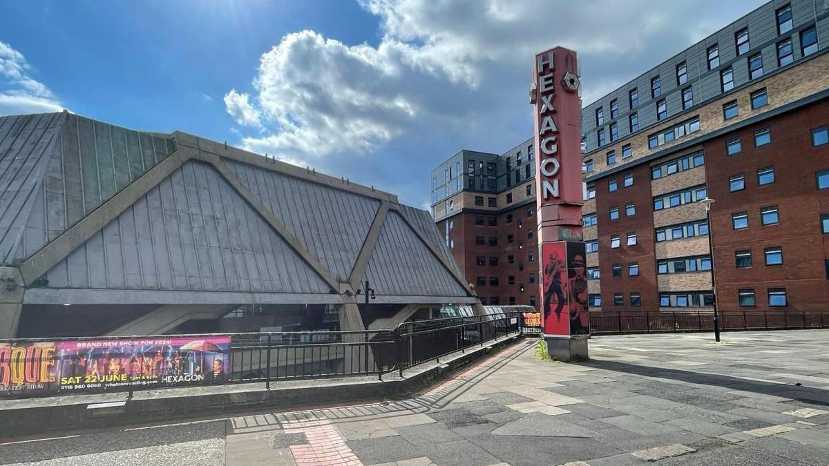 A photo of The Hexagon as it is now on a sunny day. The pillar saying "The Hexagon" is on the right, and the building is a rectangular grey building with a block of flats behind it.