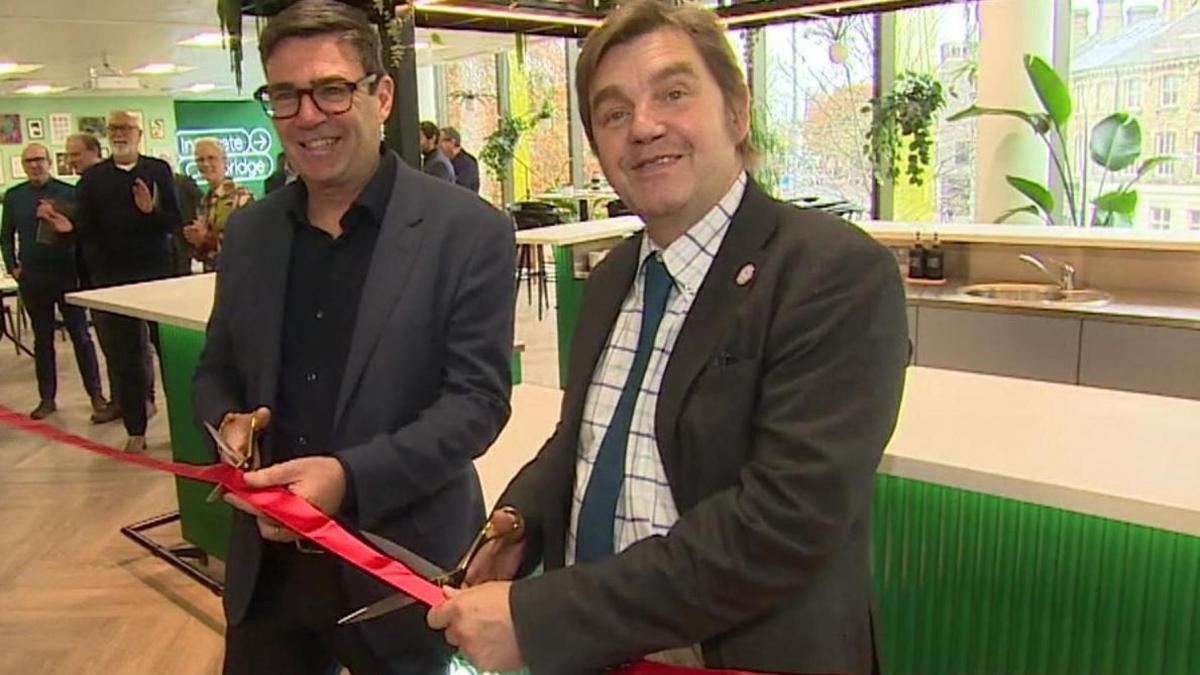 Andy Burnham stands to the left, dressed in a dark suit, with Nik Johnson, to the right, in a white and blue checked shirt and dark grey suit. They each hold a pair of scissors to cut the red ribbon.