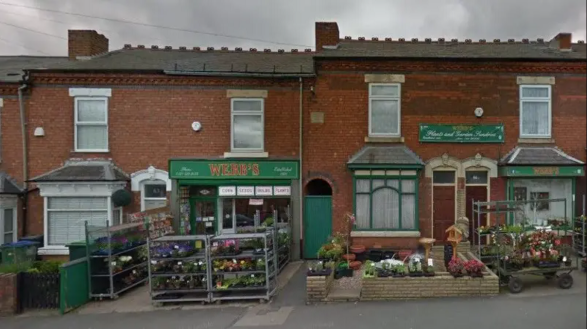 The front of a shop, which has green signage above the door and racks of flowers and plants outside. It is situated among a row of terraced houses.
