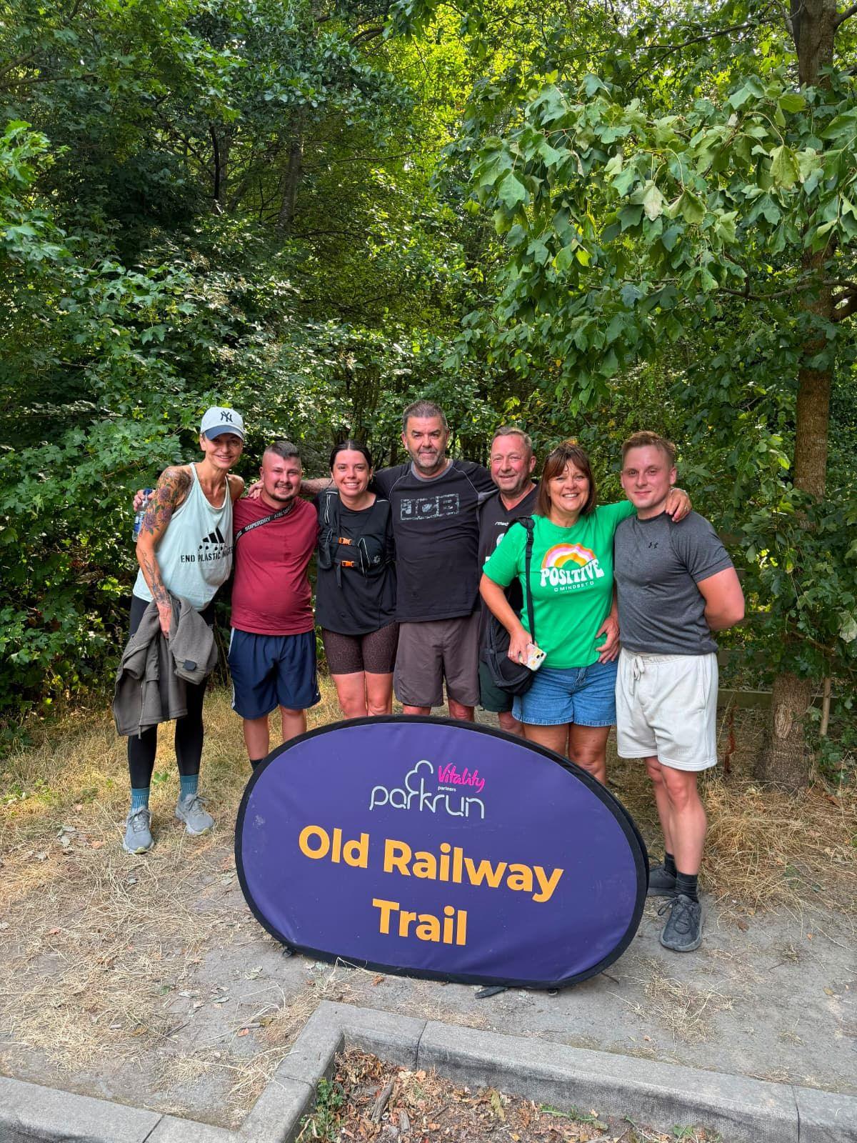 Seven people stood behind a parkrun-branded purple and orange sign which reads: Old Railway Trail. They have their arms around each other and are smiling and looking at the camera. Behind them are trees and bushes.