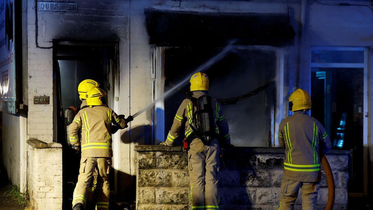 Firefighters working to put out a fire in a house during the riots. They are wearing their uniform with yellow helmets. There are four of them in the picture.