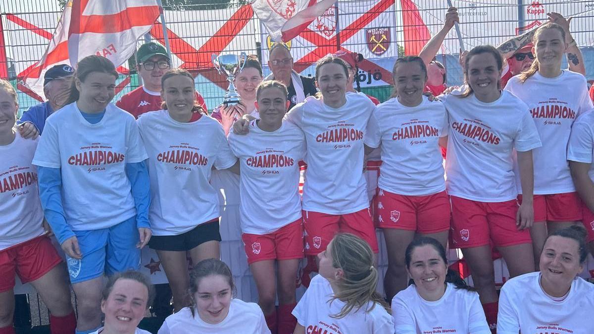 Jersey Bulls Women celebrating winning the title by wearing white t-shirts with champions on it while a trophy is held up behind them.