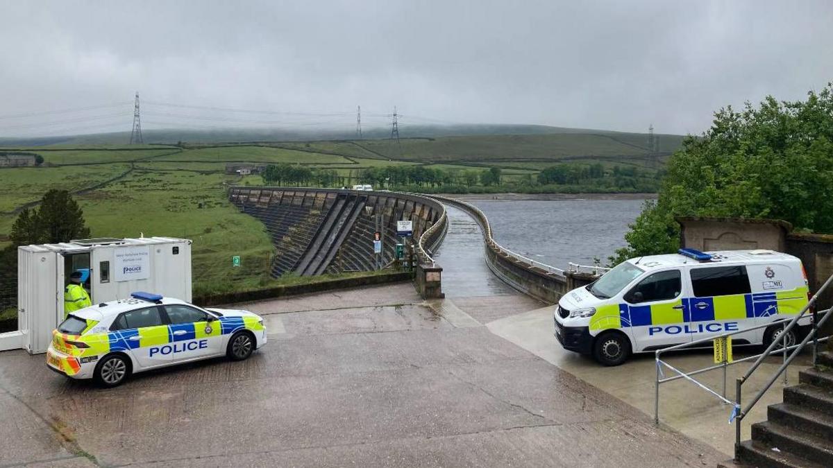 Police vehicles in the foreground, with a dam and reservoir in the background.