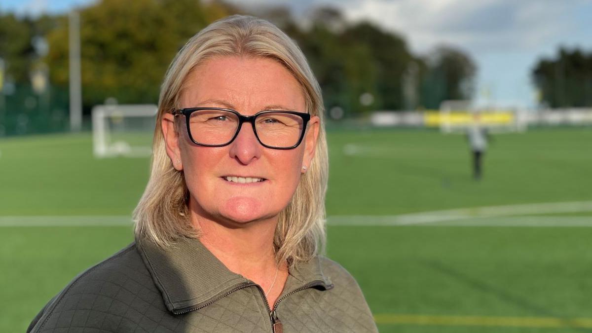 A head and shoulders photo of Jackie Thornton who has shoulder length blonde hair and black glasses, is stood on a football pitch