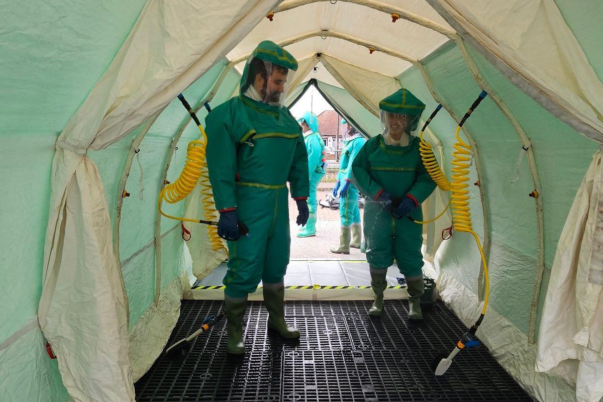 Two hospital staff wearing green hazmat suits are in a decontamination shelter, or tunnel. They appear to be smiling. Other staff in hazmat suits are in the background outside of the shelter.