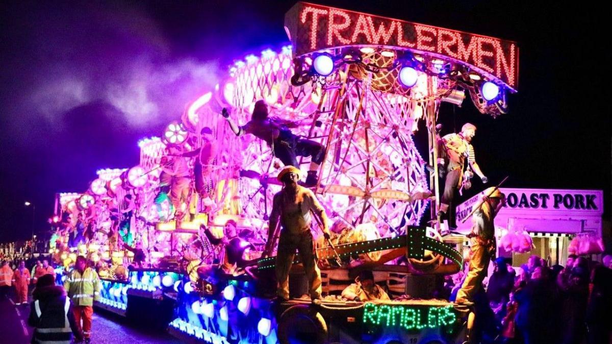 A lit up carnival cart at Bridgwater carnival in 2023. The lights are pruple, red and blue. There are dressed up people standing on it and a crowd around the cart.