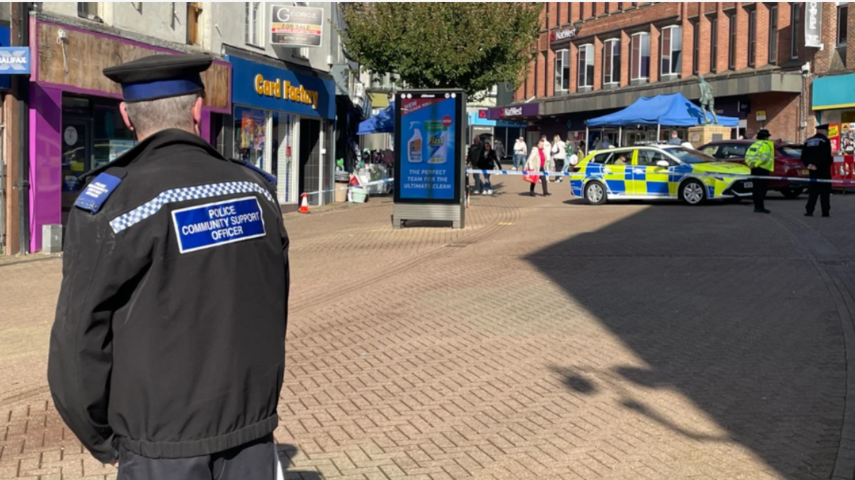 Shops and buildings can be seen either side of the main pedestrian route on Parliament Row. A police car and cordon is visible and shoppers stop to look at the scene. A police officer is in the foreground watching the scene.
