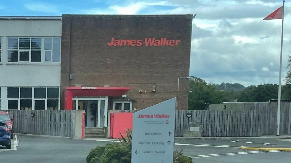 Photo from the car park of James Walker building. The large brick building has James Walker written in red on it. There is a sign in the car park directing cars and a flagpole in the right of the picture.