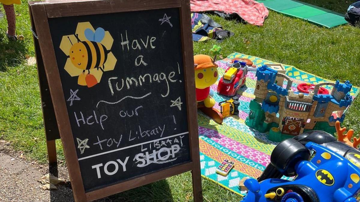 Chalk board on grass saying "have a rummage" and "help our toy library" next to rug with toys on top