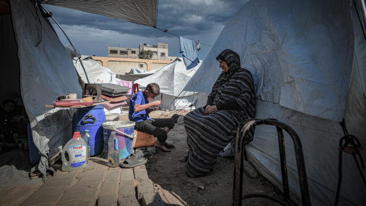 A woman and a boy sit on stools outside a tent in what appears to be a tent village. The boy is looking at something in his hans. There are a serious of buckets and storage containers scattered around them.