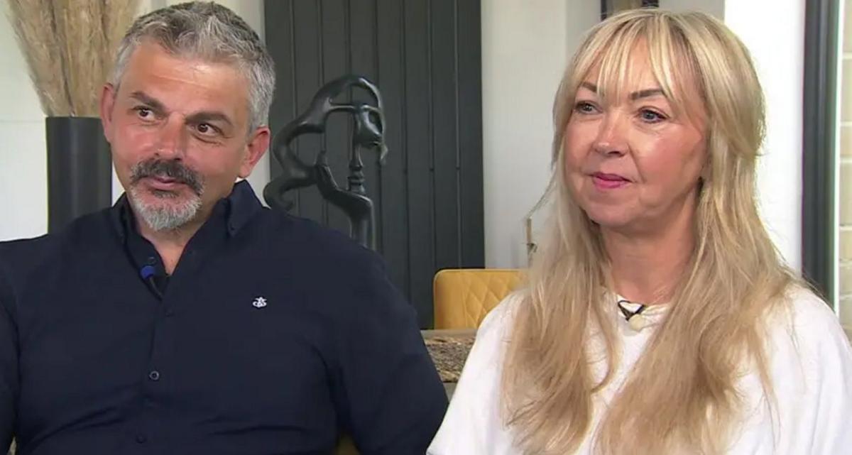 Andrew and Lisa Roussos sitting side by side at home. Andrew is wearing a black shirt, has short great hair and a goatee beard and Lisa is wearing a white top and has long blonde hair