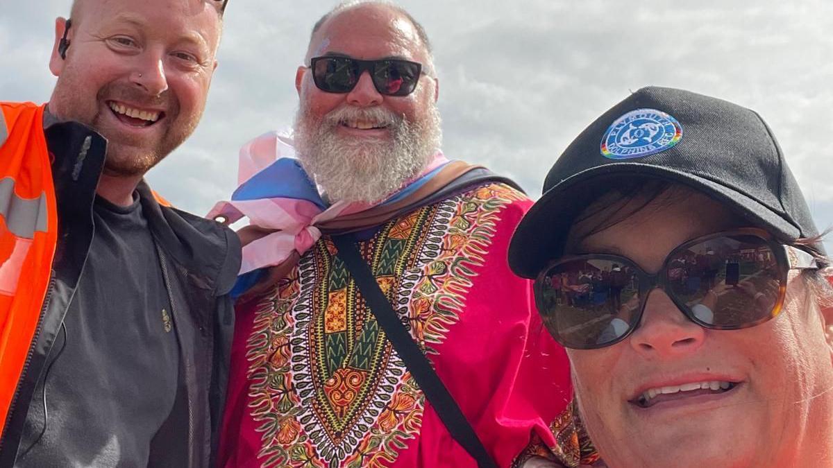 Alex is pictured with two other volunteers outside at a previous Plymouth Pride event. All three are smiling at the camera, while Alex wears an orange hi-vis jacket over a dark top.