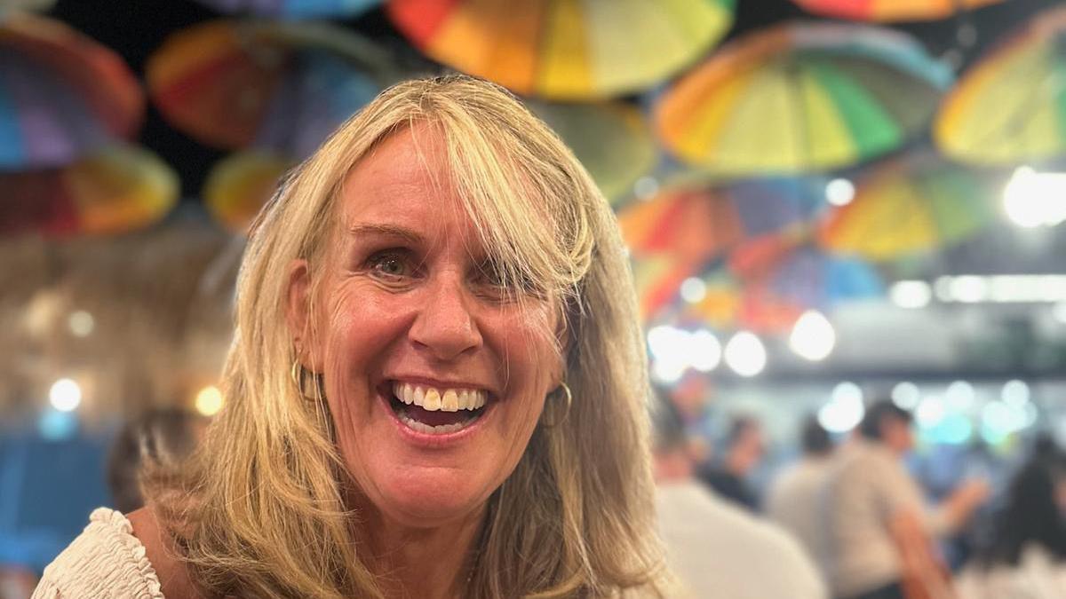 A picture of Jane Cave. She has a large smile on her face and is wearing a white item of clothing which can be just seen on her shoulders. She has blonde hair and is wearing gold earrings. In the background decorative colourful umbrellas can be seen on the ceiling behind her. 