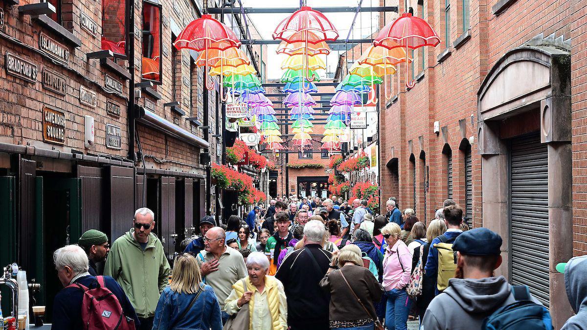 A crowd of people. They are walking down a narrow street with red-brick buildings on either side. Above them are fake umbrellas of various colour.