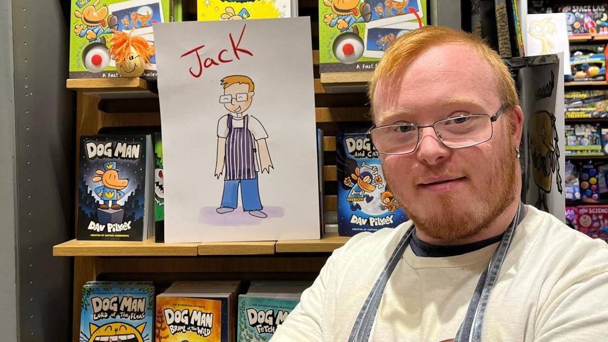 Jack Dillon smiles at the camera next to an illustration of himself from the book. He has short ginger hair and a beard, blue eyes and is wearing glasses, a white shirt and a blue apron.