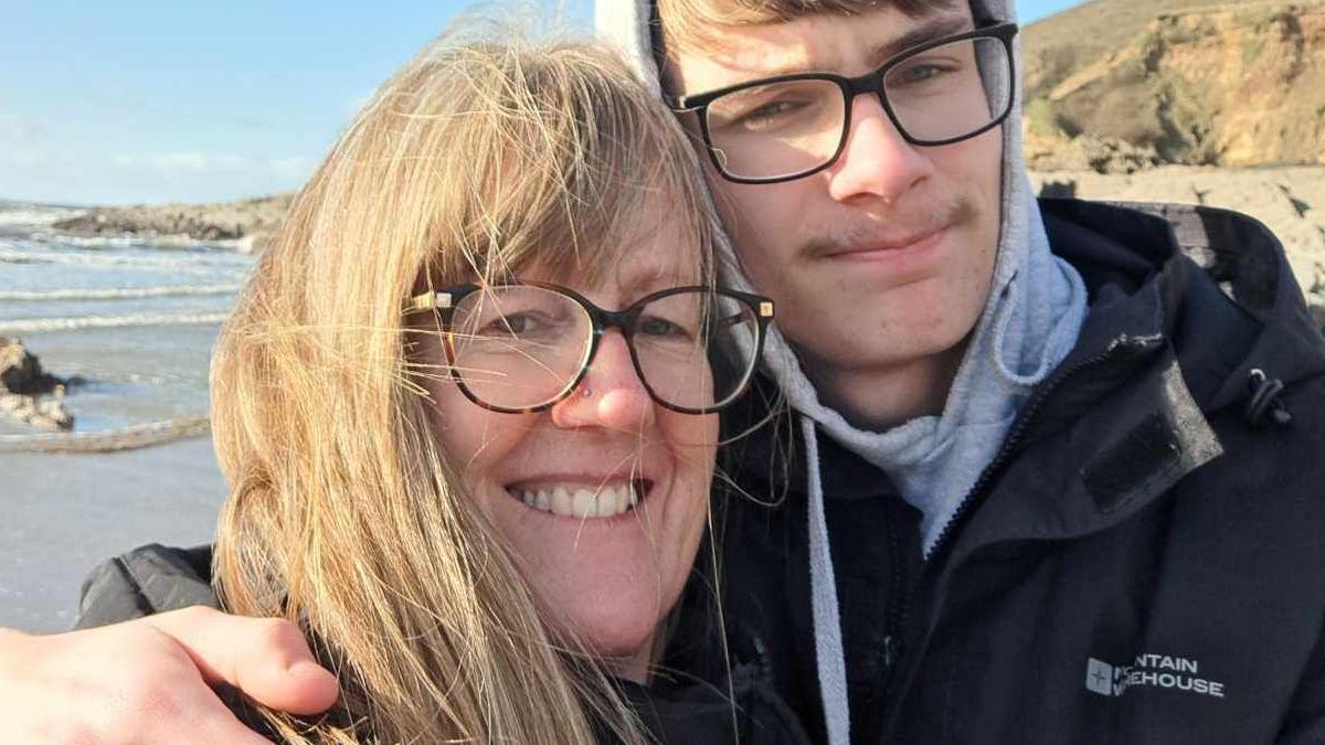 Sally Donley, a blonde woman with glasses, is being hugged by her son Euan, who is wearing glasses and a grey hoodie under a black jacket. They are pictured on a beach, with the sea and cliffs in the background. Both are smiling at the camera.