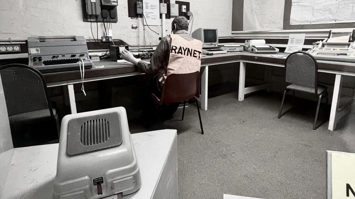 Rear view of a man in a hi-vis vest sitting at a row of desks full of 1980s computer and telecoms equipment and machinery.