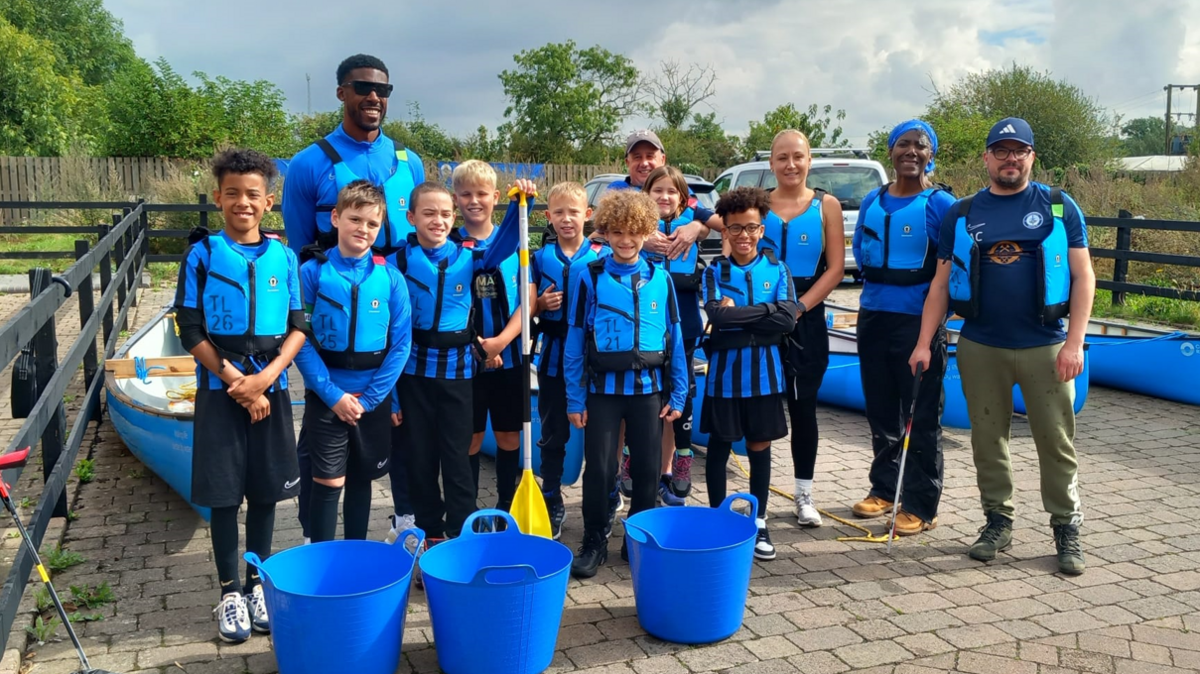 A group of children and adults wearing blue football and training kits and life jackets with canoe paddles and large containers.  They are standing in front of a row of canoes