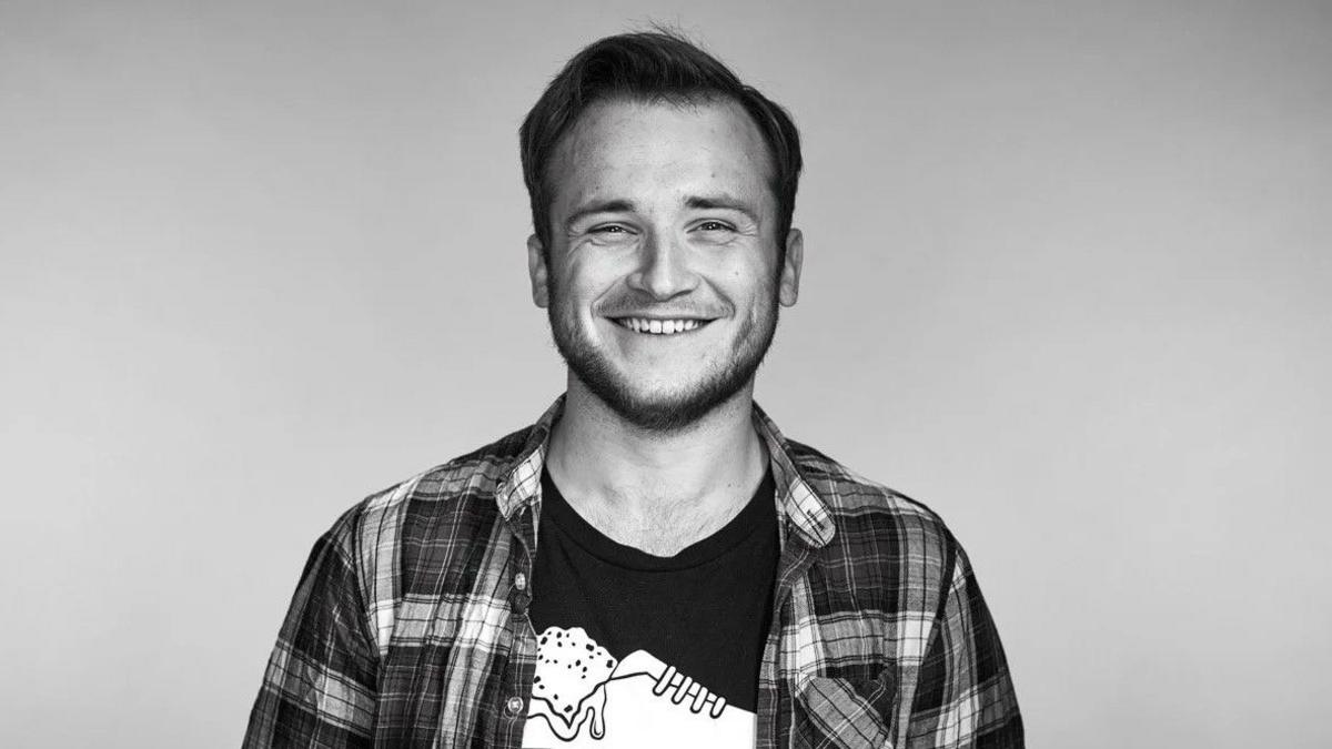 A black and white photograph of Joseph Harrington, who is looking directly at the camera and smiling. He is wearing a checked shirt over a T-shirt, featuring a training shoe design. 