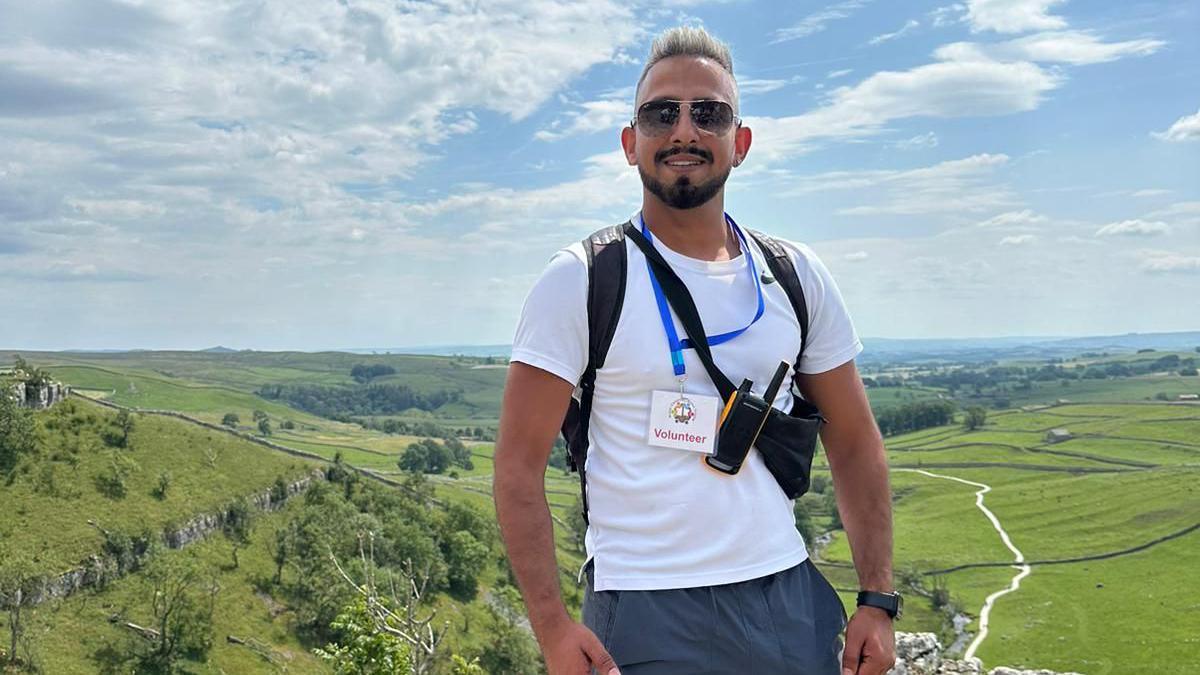 A young man stands in a beautiful countryside setting, possibly on top of a hill, with rolling hills and fields behind him. He wears a white t-shirt, and round his neck is a lanyard with a 'volunteer' badge in it. He also has a walkie-talkie attached to his side.