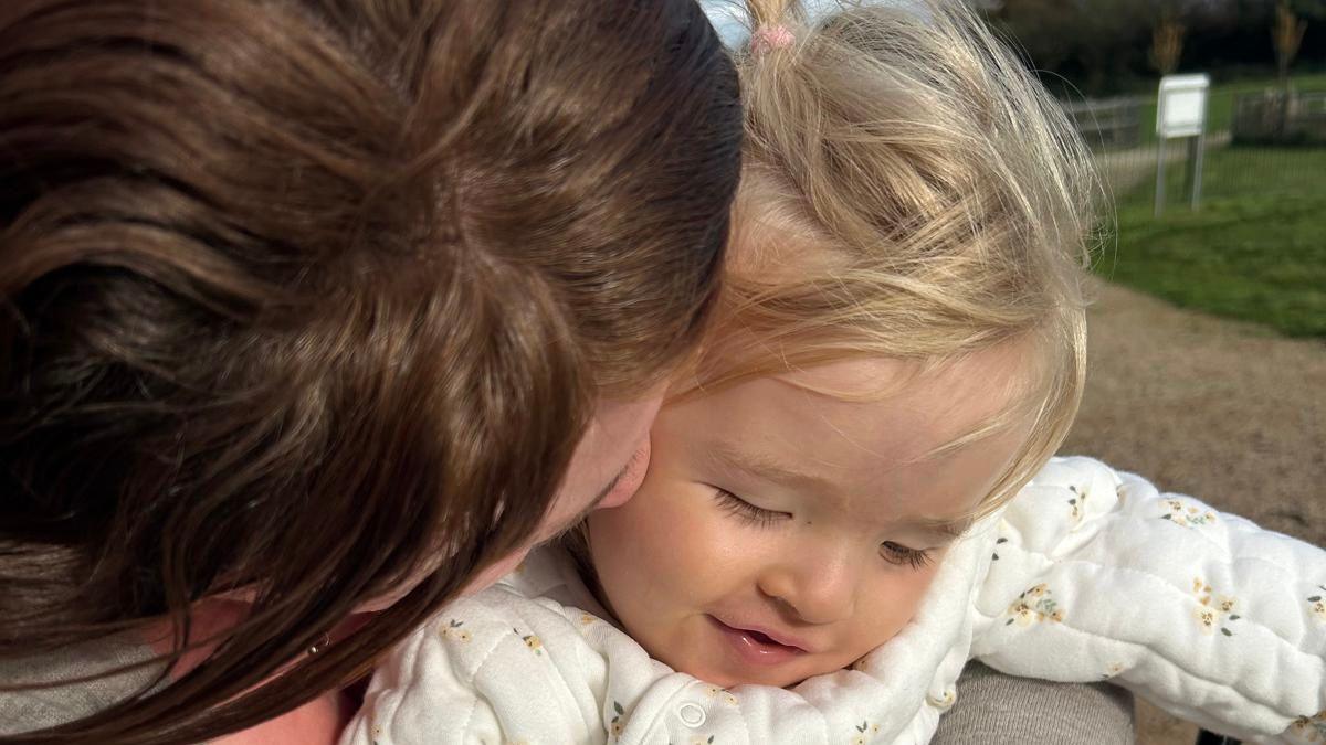 A baby with blonde hair in a white jumpsuit. She is being embraced by her mother with brown hair. 