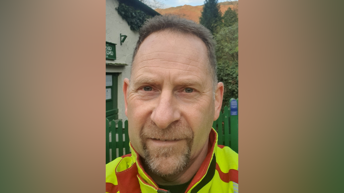 Neil Aitken has short brown hair and facial hair. He is wearing a fluorescent yellow and red firefighters uniform.