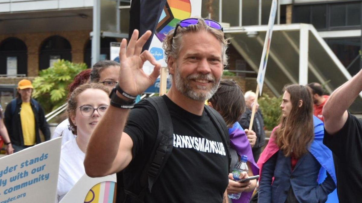 Mark Harvey doing an OK symbol with his thumb and forefinger on his right hand, as he smiles at the camera, while wearing an all black t-shirt with "Andy's Man Club" emblazoned on the front of it. He appears to be in a crowd of people who are holding banners and walking.