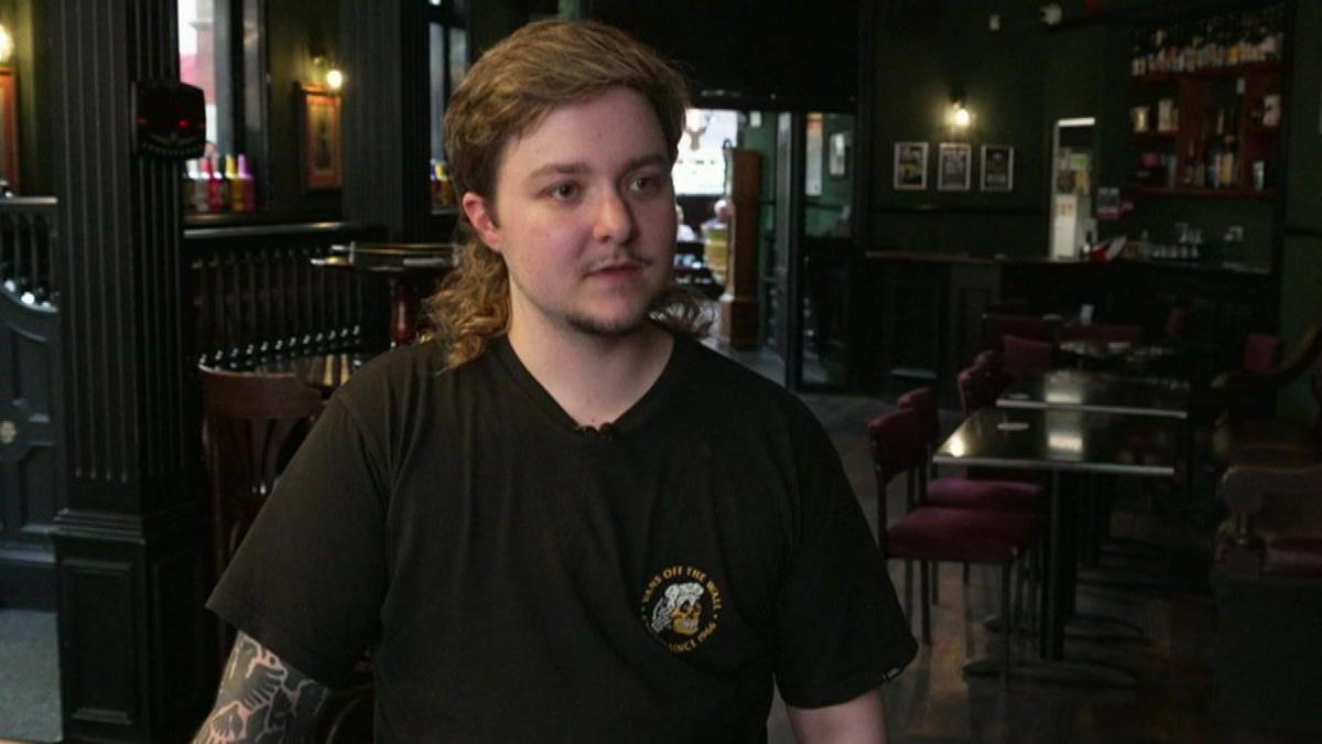 The manager wears a black t-shirt with a logo on it. He stands in the bar area which is painted in dark colours.  It is empty in the background.