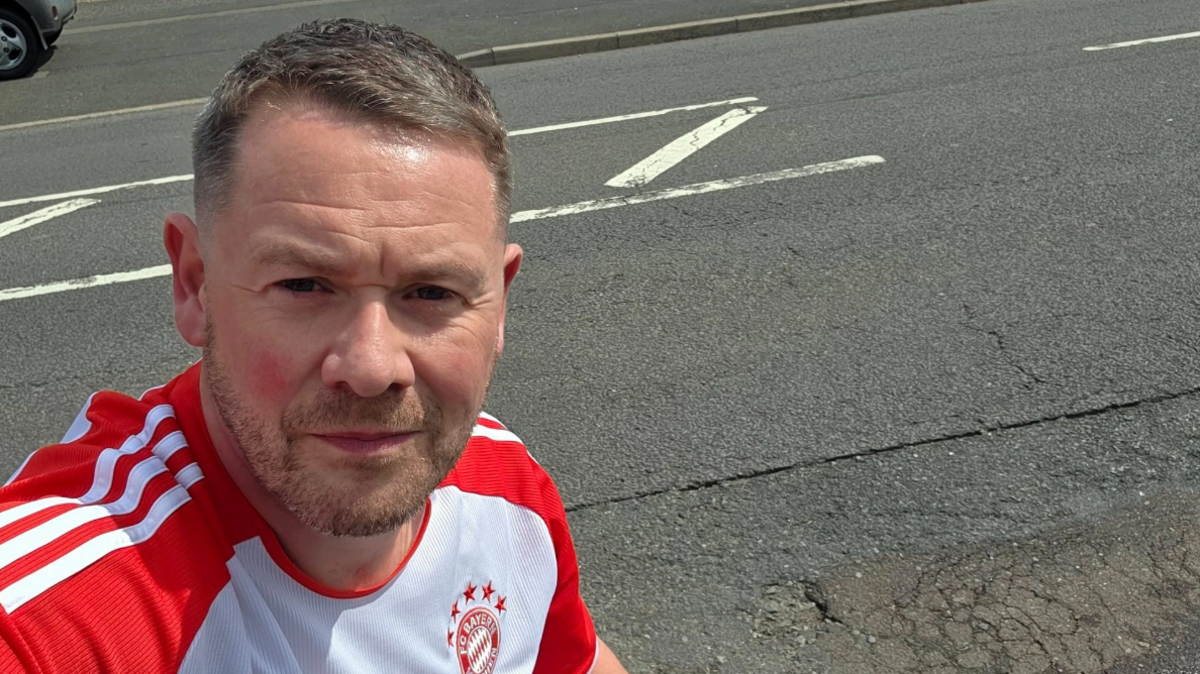 A man with a red and white FC Bayern top looking into the camera next to a road.