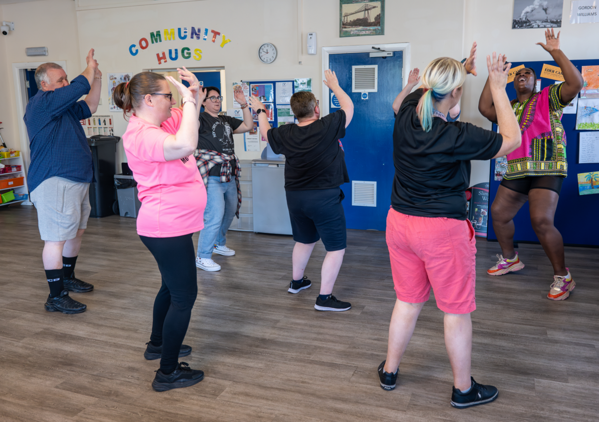 A community dance class. Five people are facing a dance instructor and copying a dance move where their hands are up in the air.