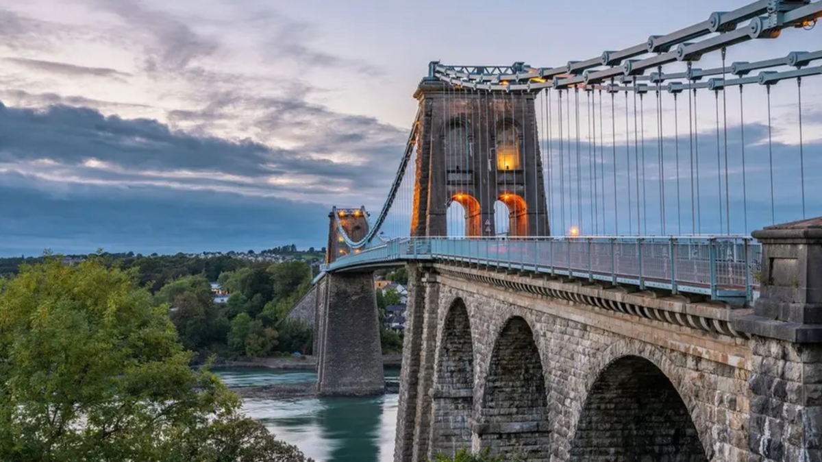 The Menai Bridge is made of stone, with blue water flowing beneath. Green trees to the left hand side. A cloud sunset in the background.