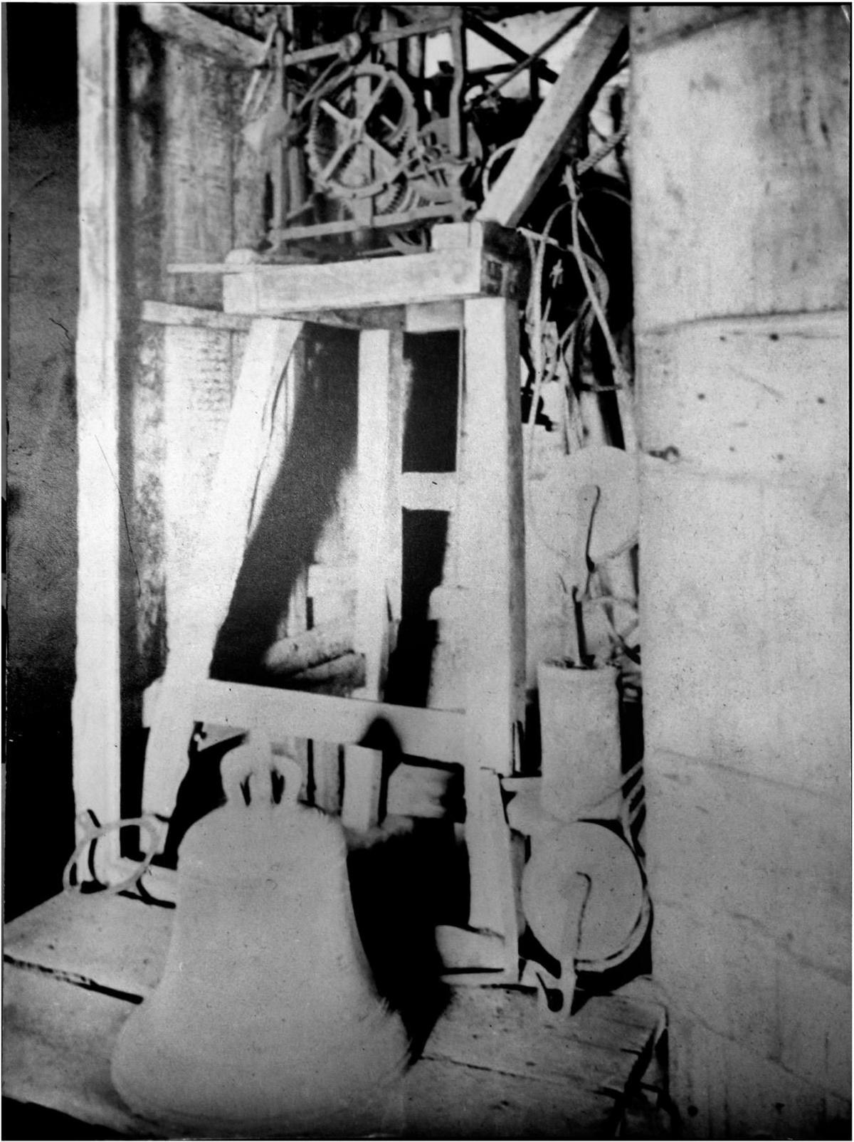 A black and white photograph shows the clock in pace with in the tower of the church with an bell unattached on the floor next to the bell frame.