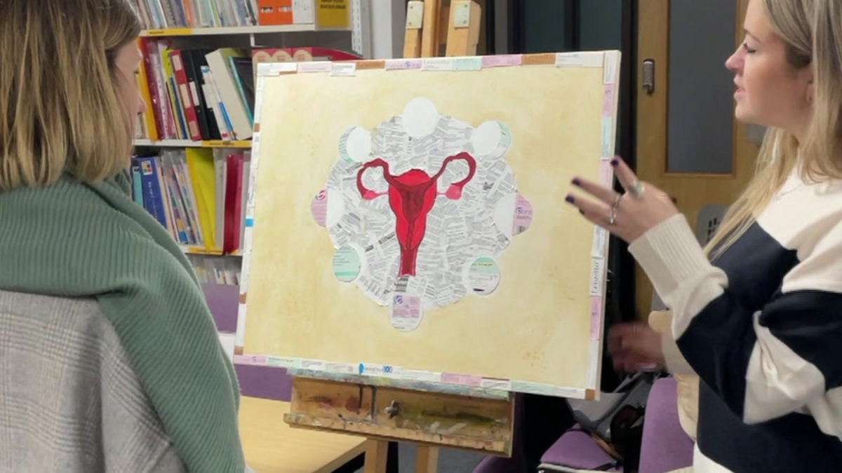 Two fair haired women discuss an artwork on an easel in front of them. The artwork has a yellow background and features a red uterus.