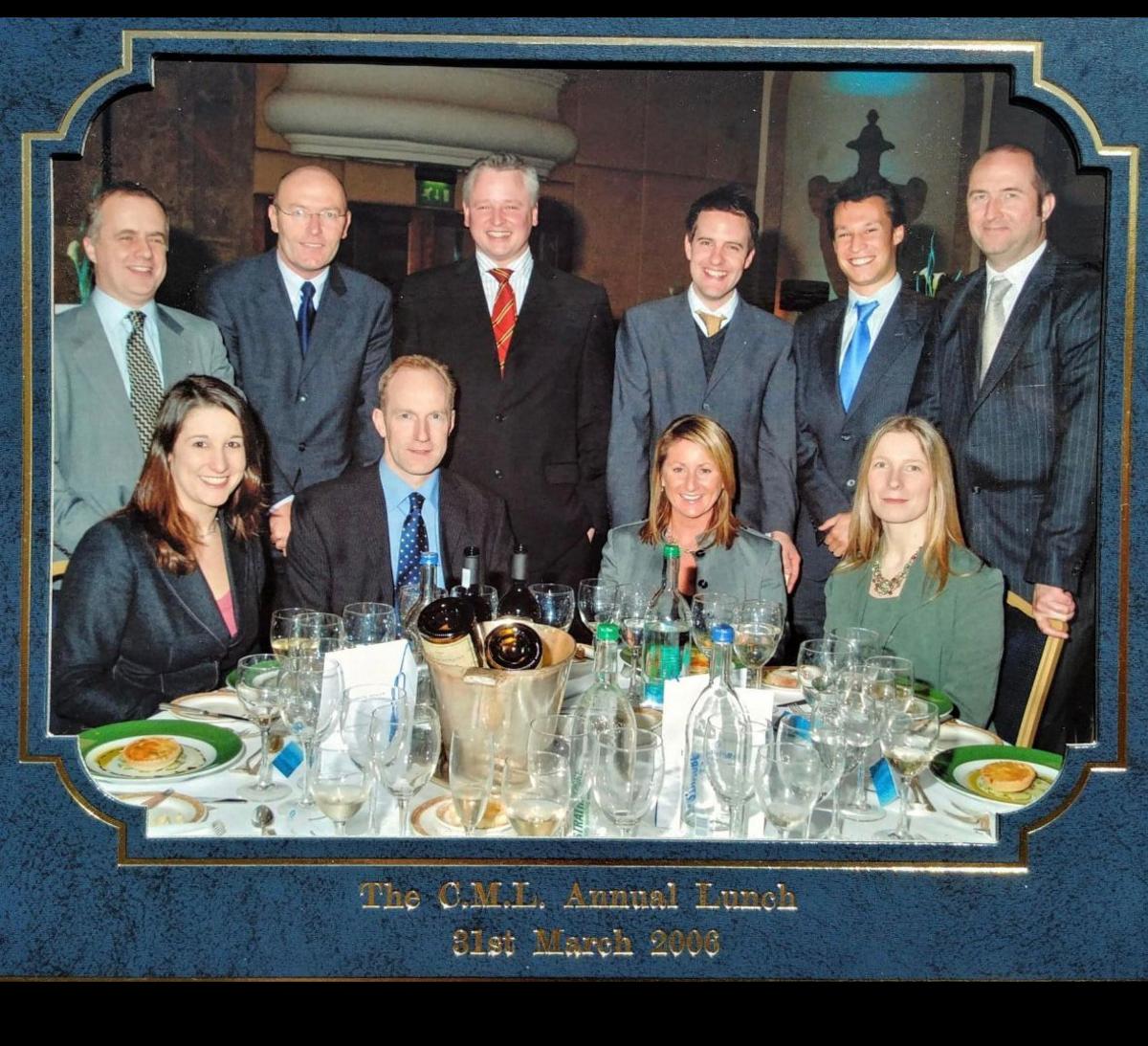 Picture of Rachel Reeves in a group of smiling at the camera. Reeves is sitting next to a man, and there are two other women seated at a table, on which there are bottles and wine glasses. Six men stand behind the seated group in a semi circle.
